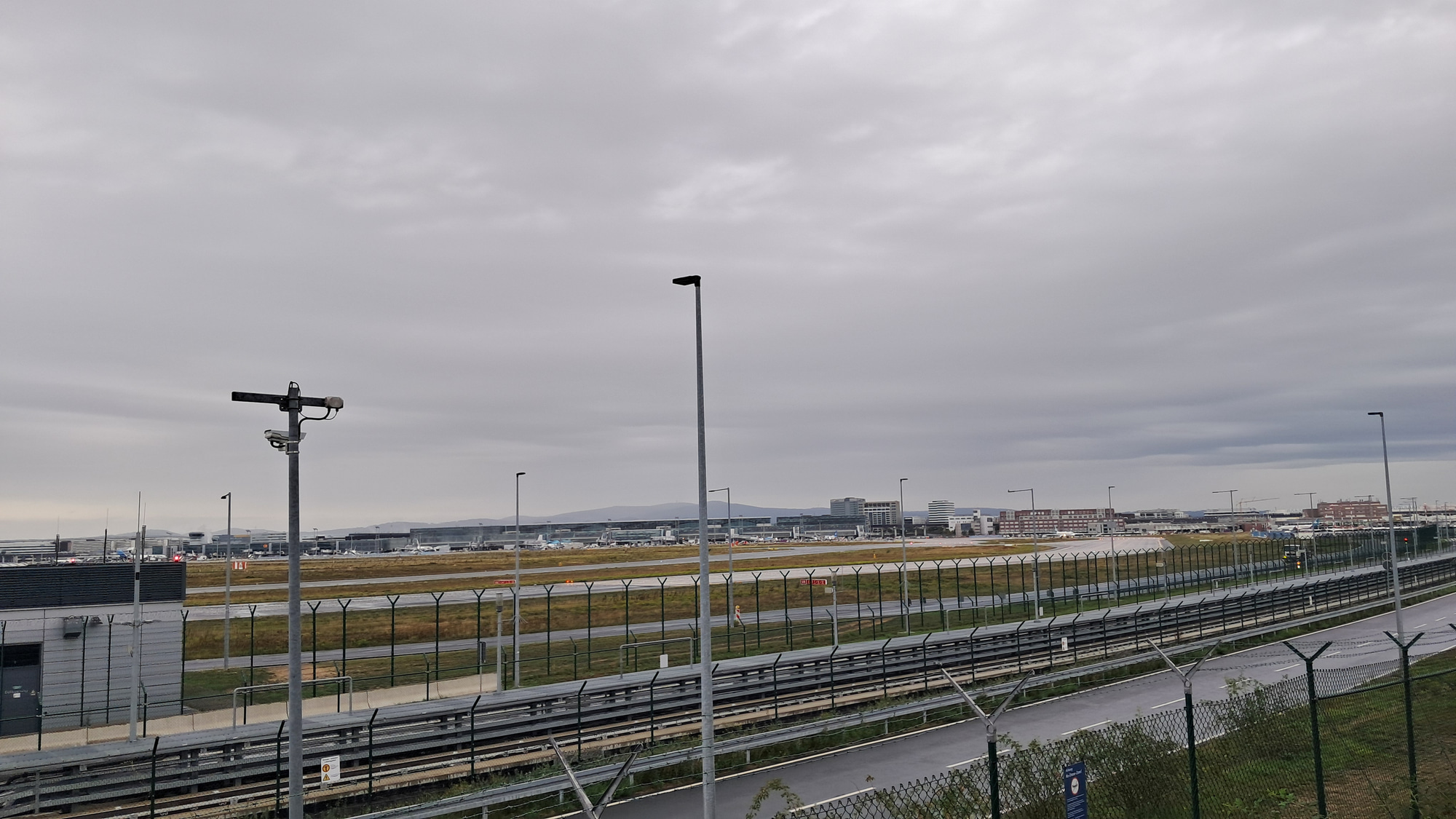 Das Bild zeigt einen Blick auf das Flughafengelände in Frankfurt am Main. Im Vordergrund befinden sich Zäune und eine belebte Straße, während im Hintergrund die Start- und Landebahnen sowie Flughafenbauten zu sehen sind. Der Himmel ist bewölkt, was dem Bild eine düstere Atmosphäre verleiht.