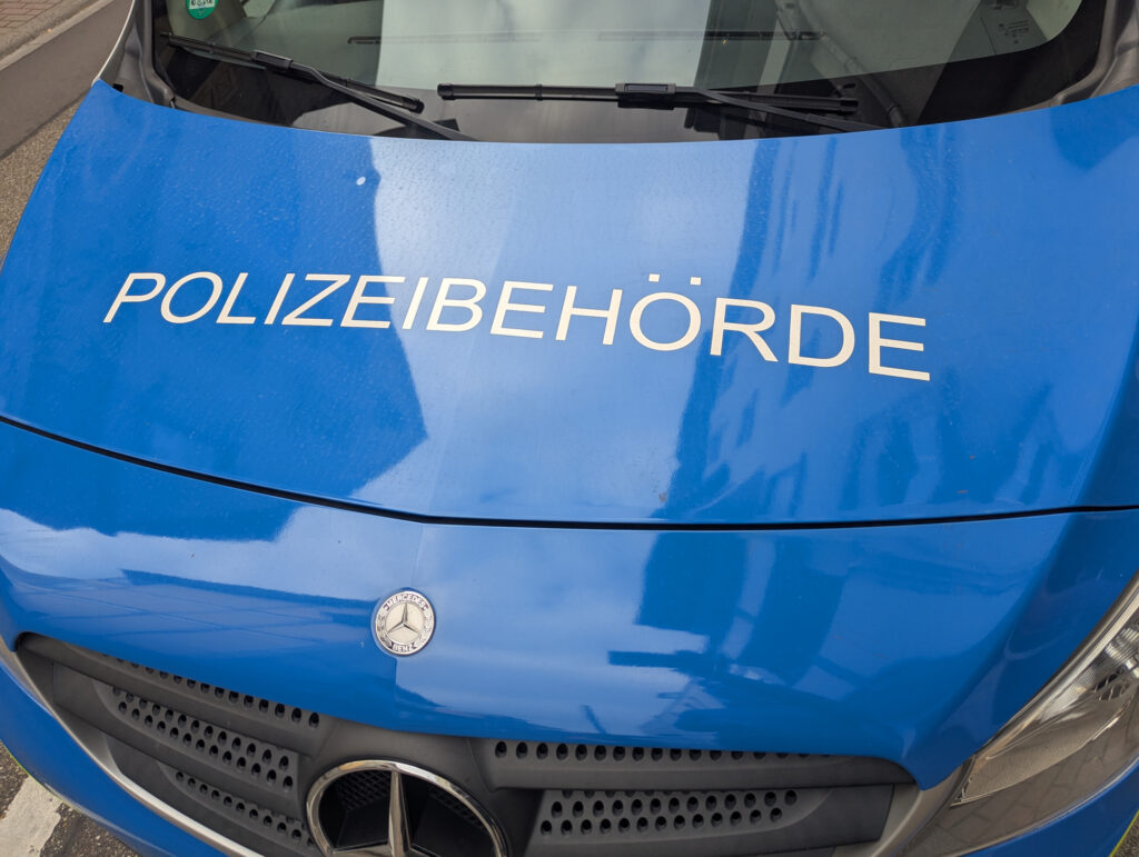 Das Bild zeigt die Motorhaube eines blauen Polizeifahrzeugs in Leimen, Baden-Württemberg. Das Logo von Mercedes ist deutlich sichtbar. Das Fahrzeug steht wahrscheinlich in einer städtischen Umgebung.