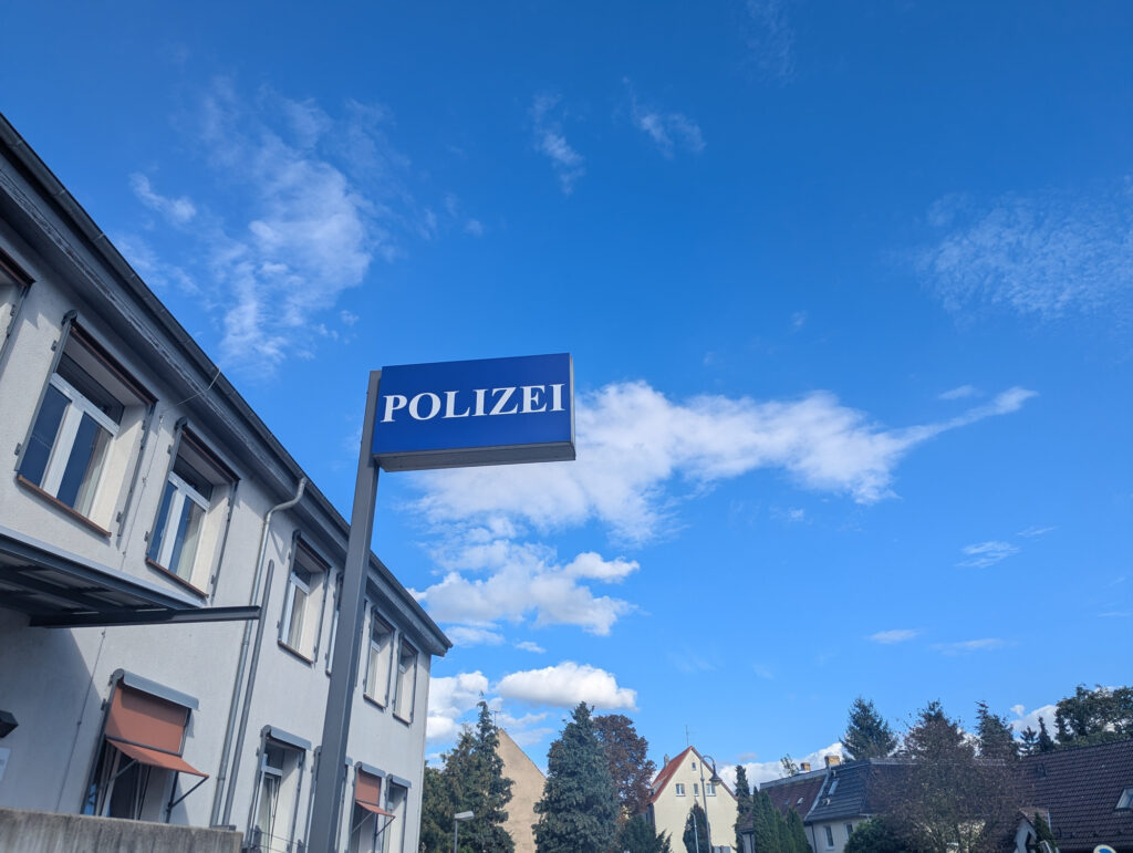 Ein Schild mit der Aufschrift Polizei steht vor einem Verwaltungsgebäude in Delitzsch, Sachsen. Der strahlend blaue Himmel mit vereinzelten Wolken bildet einen starken Kontrast zur Architektur. Das Bild vermittelt einen Eindruck von Sicherheit und Ordnung in der Stadt.