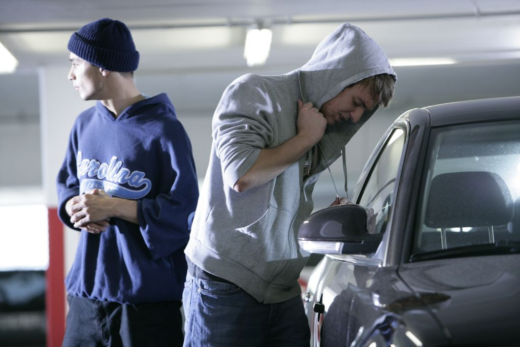 Zwei junge Männer in Kapuzenjacken stehen in einer dunklen Tiefgarage vor einem silbernen Auto. Der eine beugt sich über das Auto, der andere steht abseits.